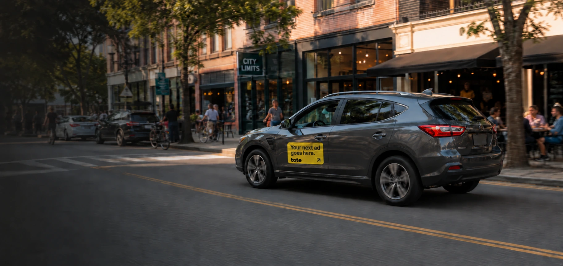 A graphite Tote campaign car passing cafes and shops on a neighborhood street